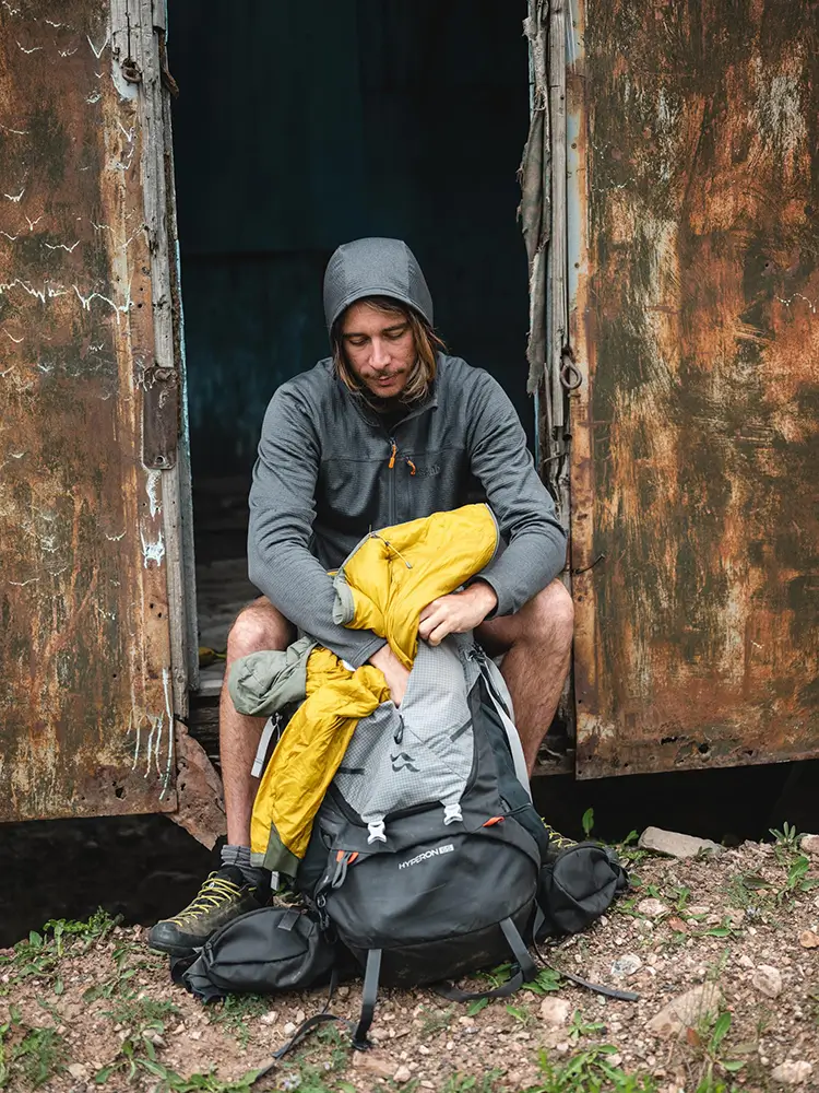 Muž v outdoorovém oblečení sedí ve dveřích opuštěné chaty a balí žlutou bundu do turistického batohu Rab Hyperon.